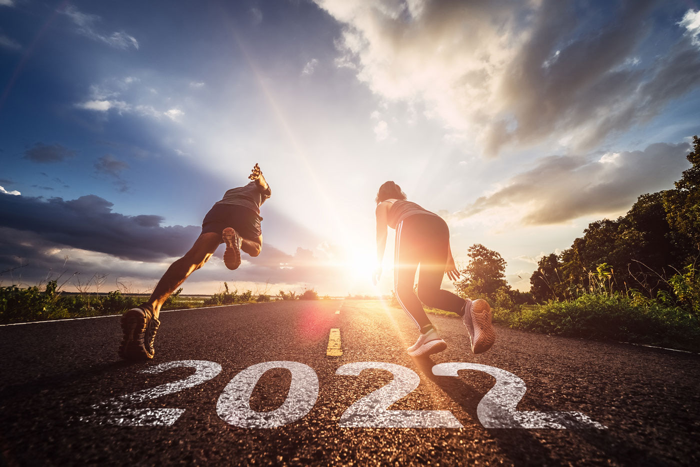 Two people in athletic wear sprint down a road at sunrise, with “2022” painted on the pavement, a dynamic perspective on New Year’s Resolutions, capturing the drive to chase new goals.