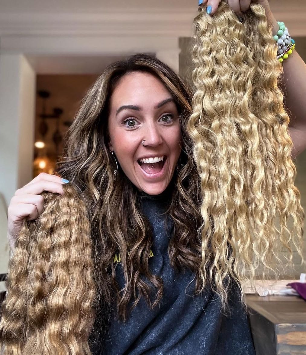 A smiling woman with wavy brown hair holds up two long, wavy blonde hand-tied hair extensions, one in each hand, inside a brightly lit room.