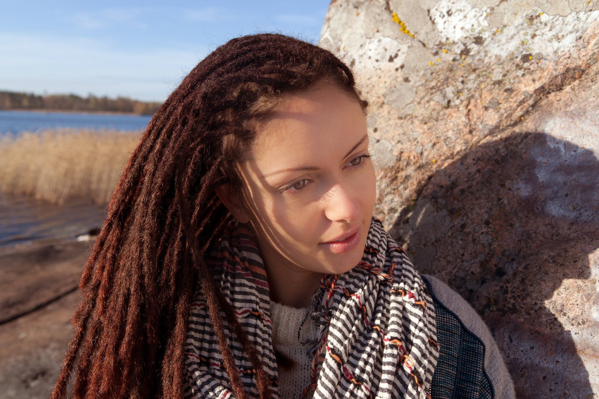 A woman with long, brown dreadlocks and a striped scarf stands outdoors near a large rock, water, and reeds.
