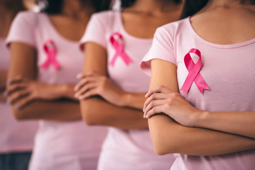 A group of women, including Priyanka, stand with arms crossed in light pink shirts and pink ribbons pinned to their chests, symbolizing strong support for breast cancer awareness.