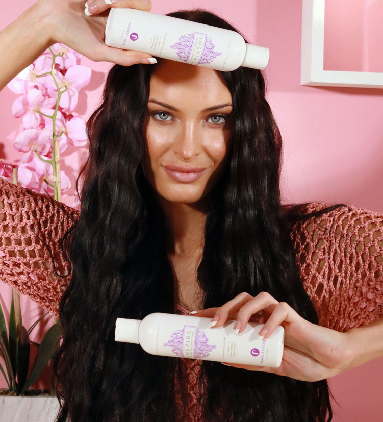 A woman with long wavy dark hair holds two scalp cleansing hair care products, one above her head and one in front of her, standing in front of a pink background with a pink orchid.