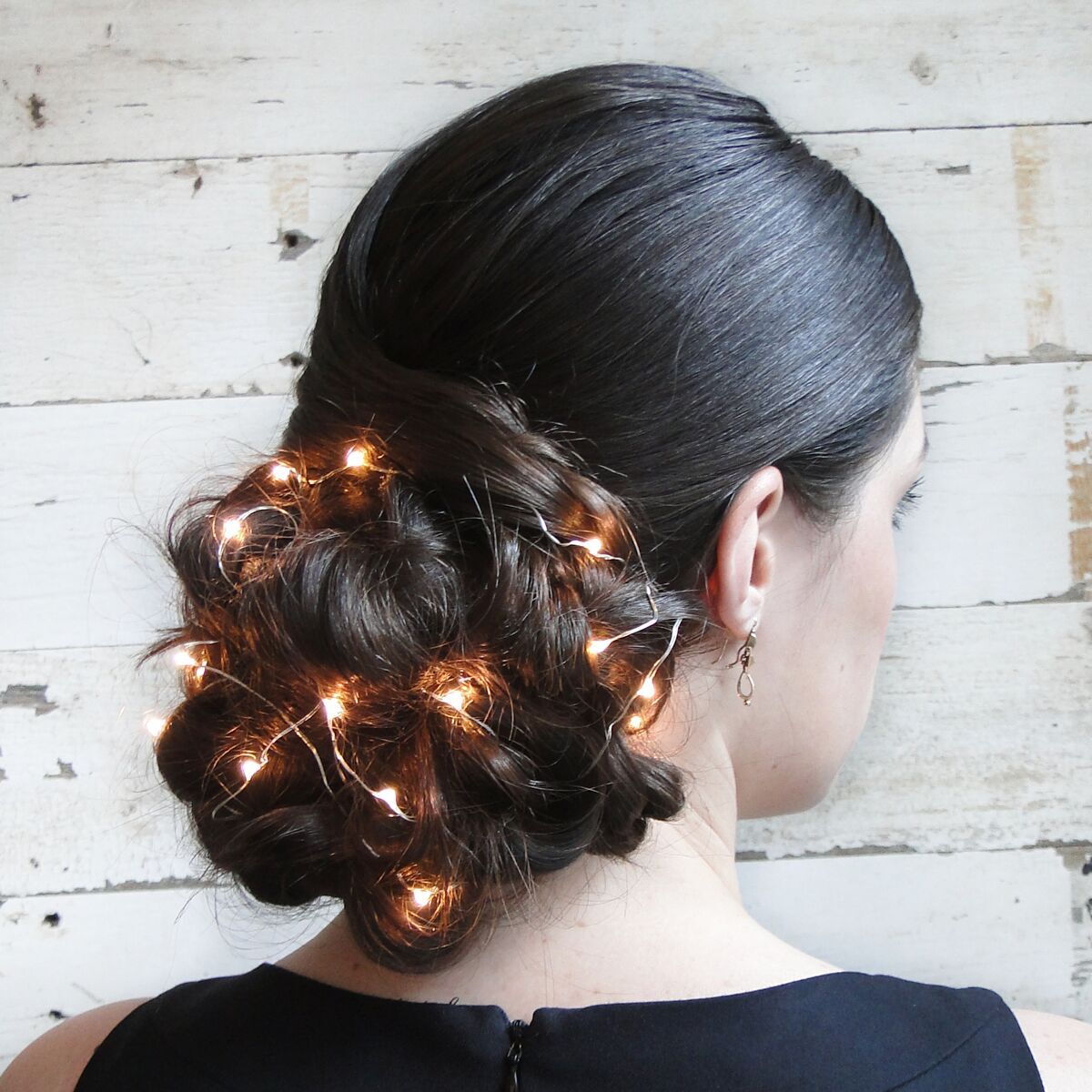 A woman with dark brown hair styled in an elegant low braided bun adorned with small, warm white string lights, wearing a black top and gold earrings.