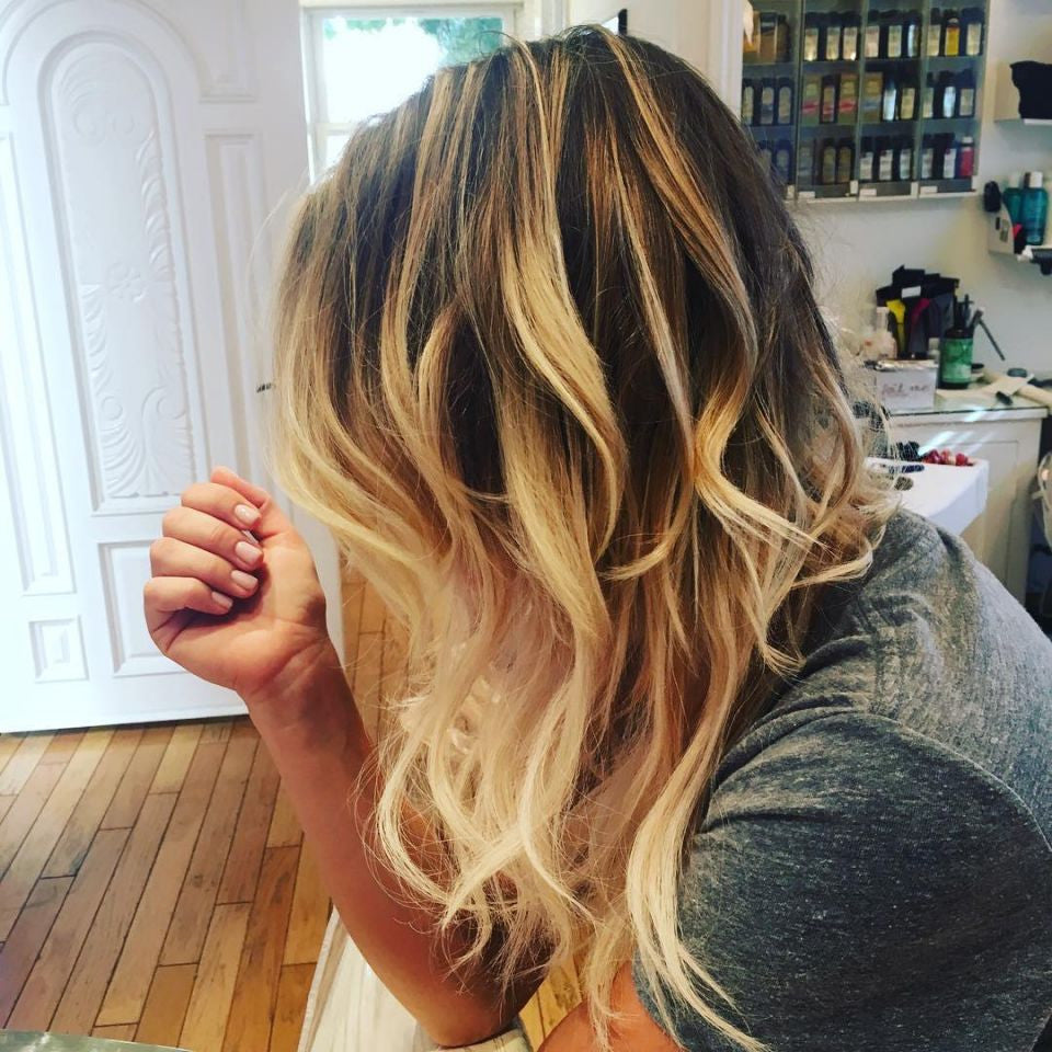 A person with wavy, shoulder-length blonde balayage hair, reflecting popular millennial hairstyles, sits in a salon wearing a grey shirt.