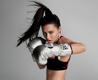 A woman with a high ponytail throws a punch while wearing metallic boxing gloves and athletic wear, showcasing gym-proof workout hair care as she looks intensely at the camera against a plain background.