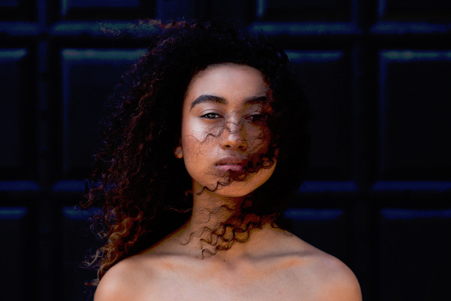 A woman with curly Virgin Indian Hair stands in front of a dark, grid-patterned background.