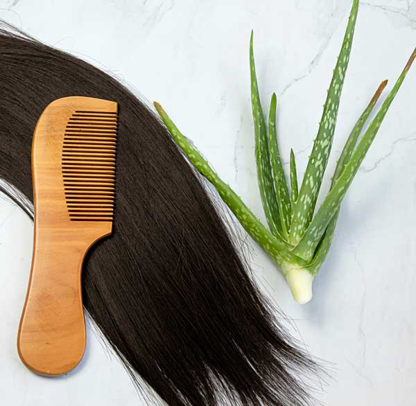 A wooden comb lies on long, smooth black hair next to a fresh aloe vera leaf, an elegant display of natural remedies for dandruff, on a white marble surface.
