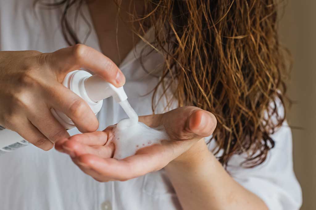 A person with wet hair, following a curly hair routine, dispenses foamy cleanser from a pump bottle into their hand while wearing a white shirt.