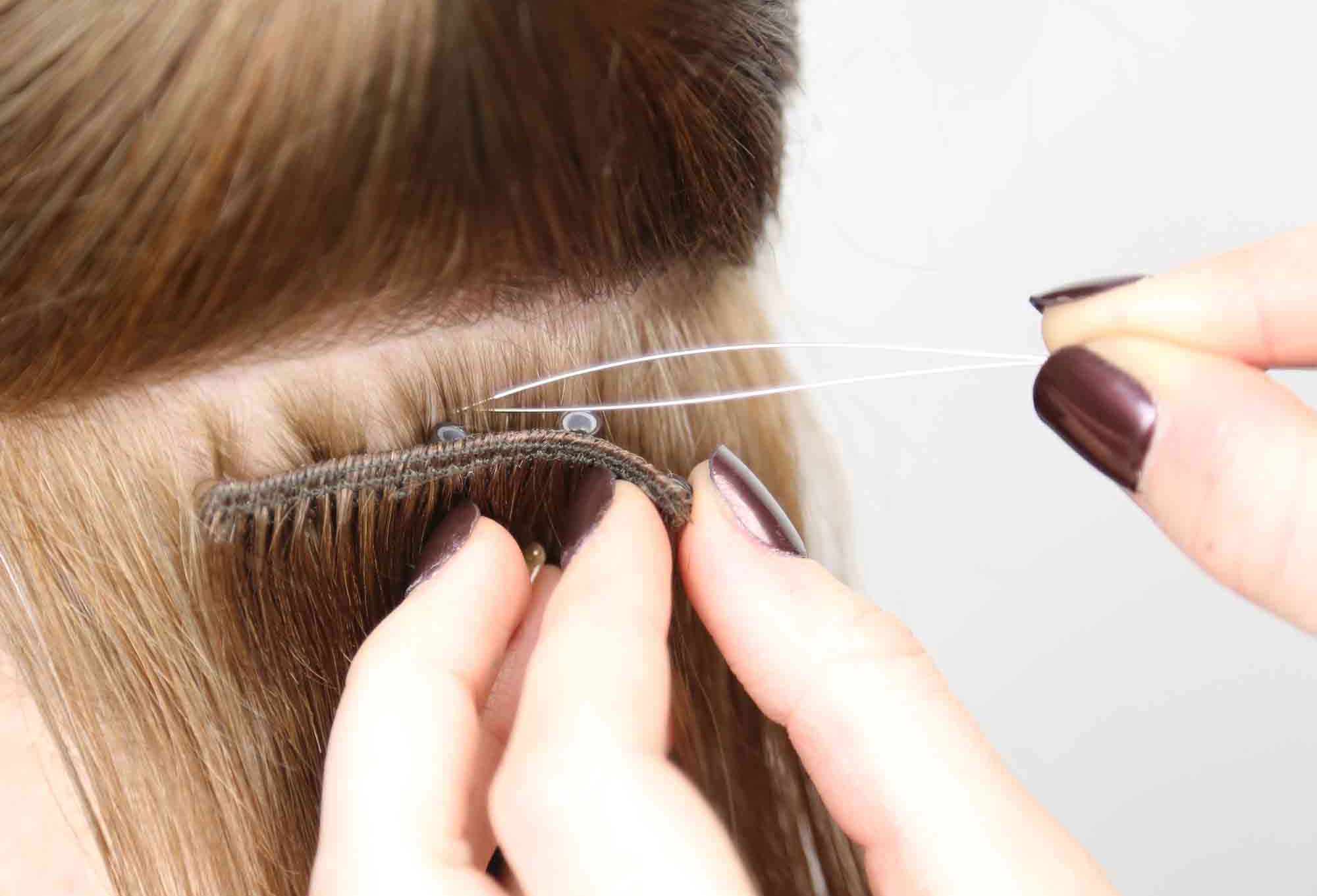 Close-up of hands attaching beaded weft extensions to light brown hair using a loop tool and small beads, with dark polished fingernails visible.