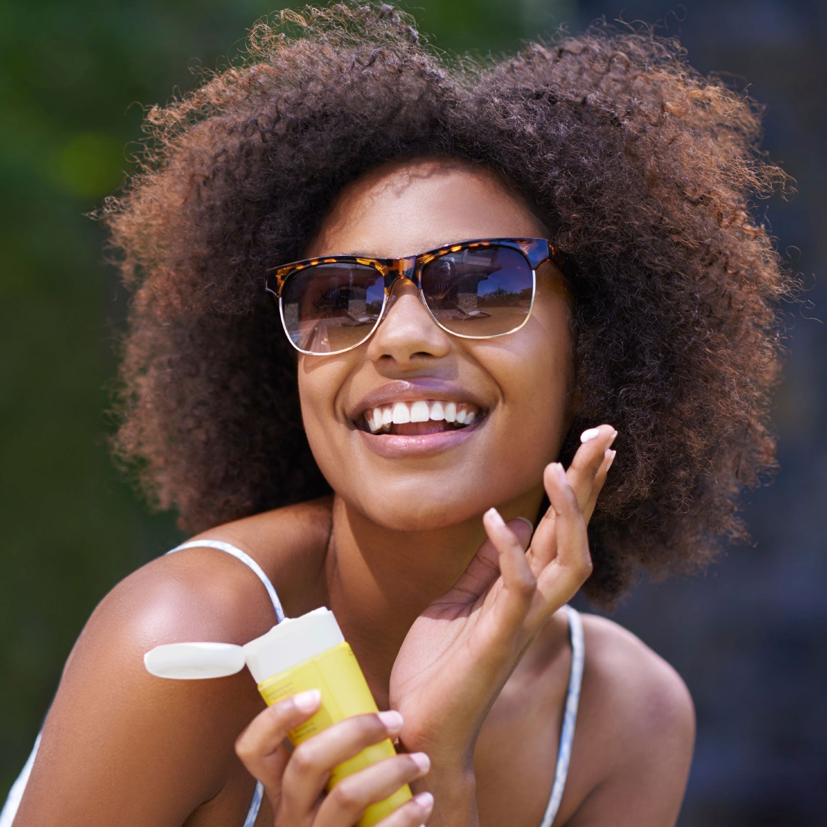 A smiling woman with curly hair wears sunglasses and a white tank top, holding a yellow sunscreen bottle outdoors with greenery in the background, ready to enjoy Labor Day while staying protected against sun and hair hazards.