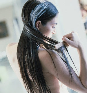 A woman with wet, dark hair applies a nourishing hair treatment for damaged hair while gently combing through it with a wide-tooth comb.