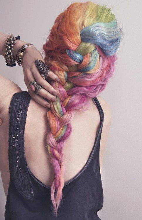 A person with long, braided hair dyed in multiple pastel colors, blue, green, yellow, orange, pink, and purple, with a homemade beauty vibe wears a sleeveless dark top and several bracelets and rings, touching the side of their head.