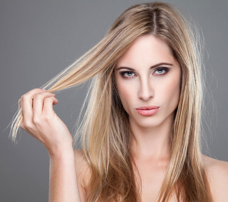 A woman with long, straight blonde hair and blue eyes looks at the camera while holding a section of her hair, showcasing hair care to get rid of flyaways.