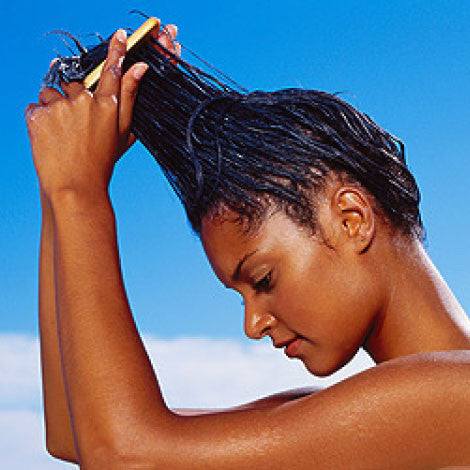 A woman practicing hair care lifts her wet hair upwards with a yellow comb under a clear blue sky, gently working to moisturize hair and keep it healthy.