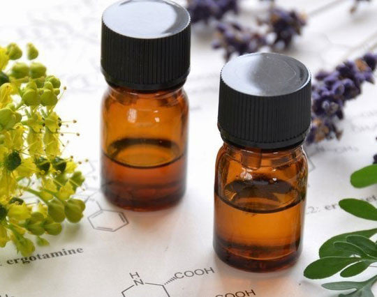 Two small amber glass bottles with black caps, filled with all-natural oils for dry hair or dry skin, rest on a paper featuring chemical structures.