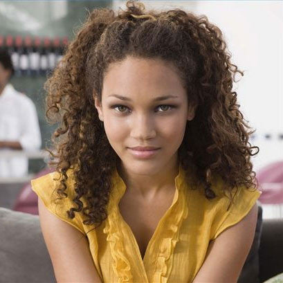 A young woman with curly brown hair, after taking her weave down, wears a yellow top and sits indoors, looking directly at the camera with a slight smile.
