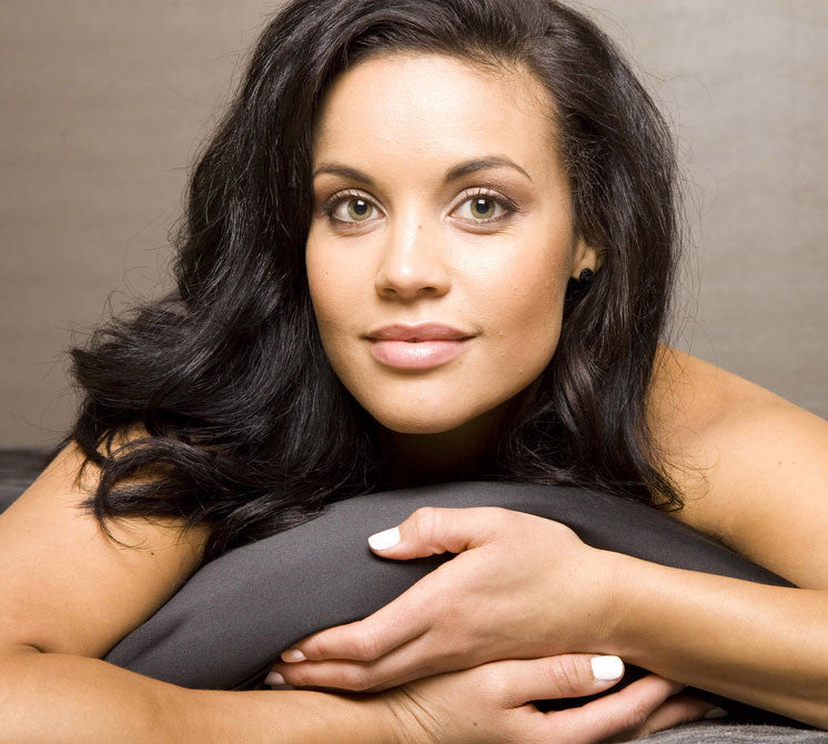 A woman with long dark hair extensions and light eyes rests her chin and hands on a gray cushion, looking directly at the camera with a relaxed, neutral expression, capturing the calm of satin dreams.