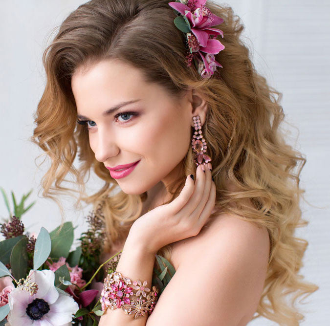 A woman with long, wavy blonde hair adorned with pink flowers holds a bouquet, showcasing perfect wedding hair.
