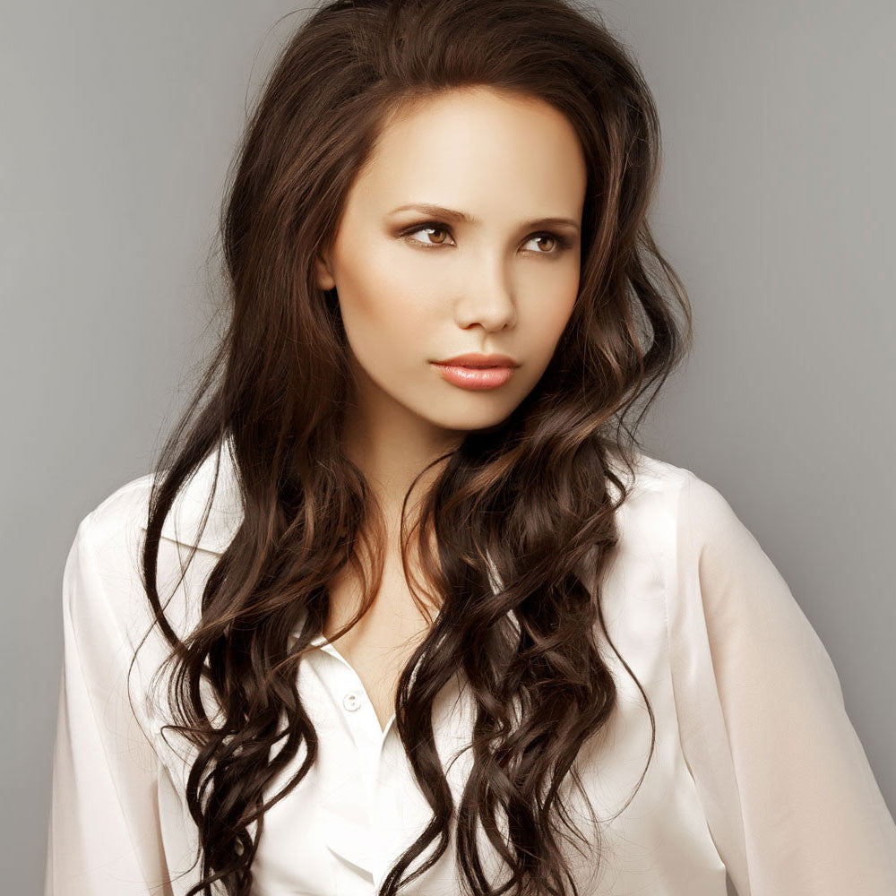 A woman with long, wet and wavy hair weave wearing a white blouse gazes to the side against a plain gray background.