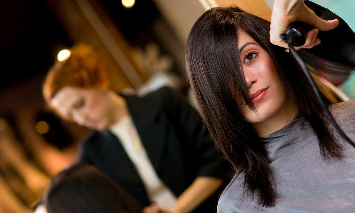 A woman with straight, dark hair sits in a salon chair, wearing a cape, while a hair stylist straightens her hair with a flat iron.