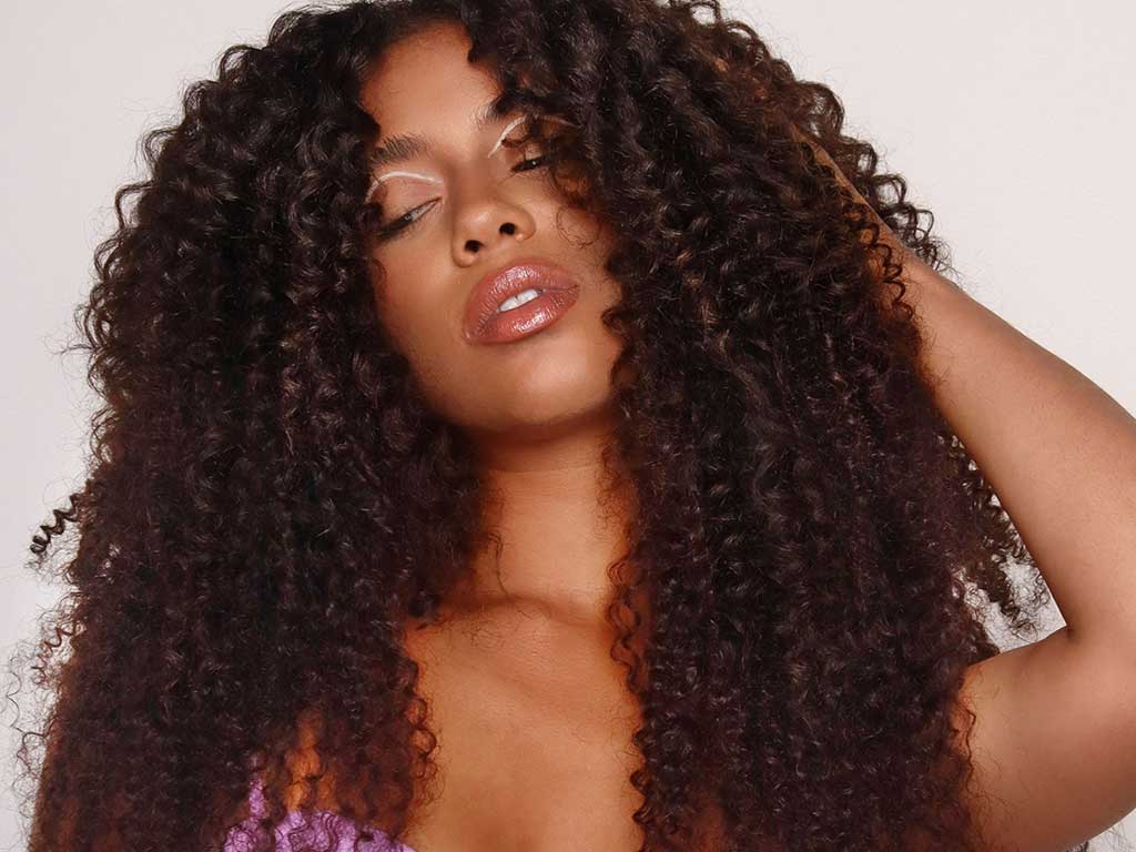 A woman with voluminous, curly hair poses with her eyes closed and one hand touching her hair, wearing a purple top and glossy makeup, her curls look nourished, as if treated with a nourishing DIY hair mask.