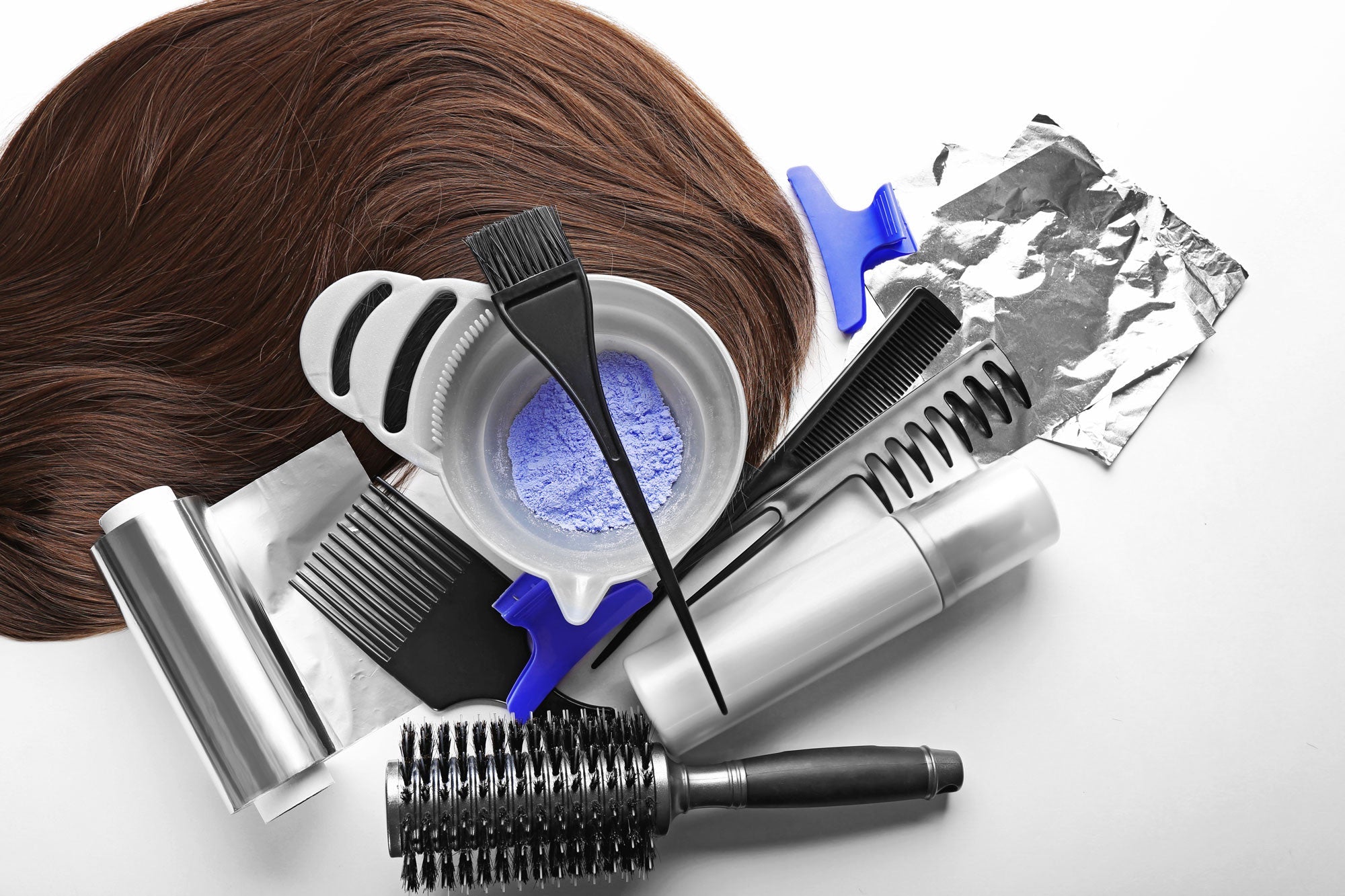Hair dye tools, perfect for those looking to dye human hair extensions, include a brush, bowl with purple powder, combs, clips, foil sheets, a spray bottle, and a round brush arranged next to a section of brown hair.