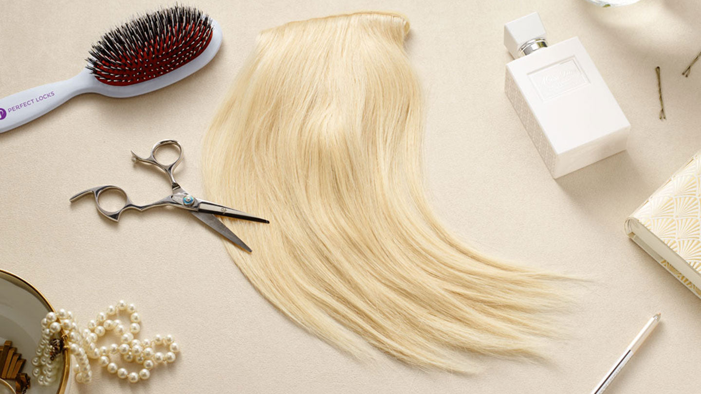 A blond hair extension lies on a beige surface surrounded by a hairbrush, scissors, perfume bottle, pearl necklace, bobby pins, and a patterned notebook.