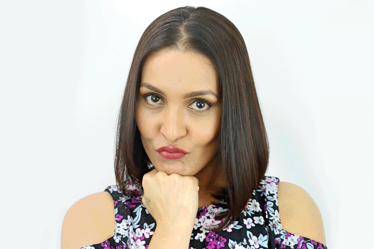 A woman with straight, shoulder-length brown hair, enhanced by a hair topper, rests her chin on her fist and makes a playful, pouty face.