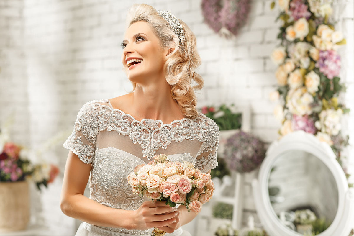 A smiling bride in a lace wedding dress showcases elegant wedding hair as she holds a bouquet of pale pink roses, standing in a bright room adorned with flowers and a round mirror in the background.