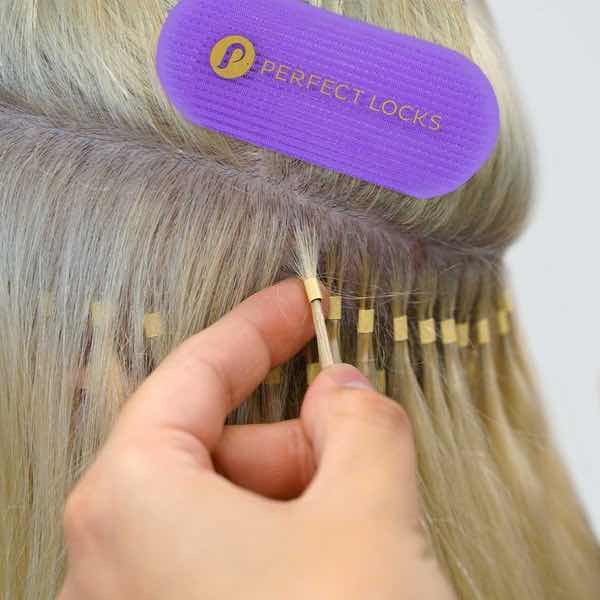 A hand installs blonde hair extensions onto natural hair using small adhesive strips, with a purple Perfect Locks sectioning clip holding hair aside, a technique among various hair extension methods, each with their own pros and cons.