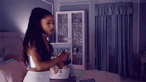 A woman in a softly lit, purple-toned bedroom puts on sunglasses and flips her long dark hair, showcasing effortless Halloween hairstyle ideas, as she turns away from an open suitcase on the bed.