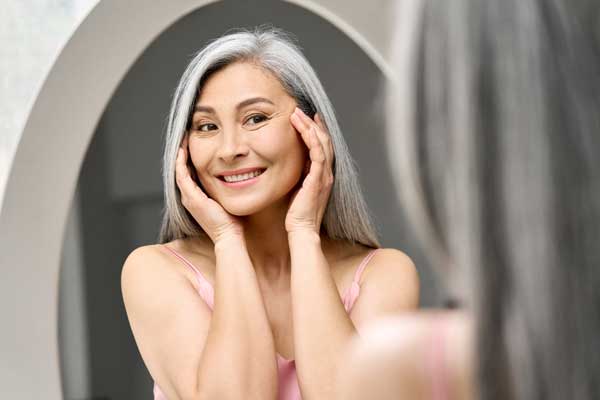 A smiling woman with long gray hair touches her face with both hands while looking at herself in a mirror.