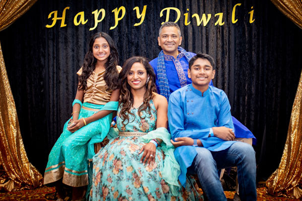 A family of four, including Priyanka, poses in colorful traditional Indian attire before a black and gold backdrop that reads Happy Diwali. Two adults sit in the center with their smiling son and daughter beside them.