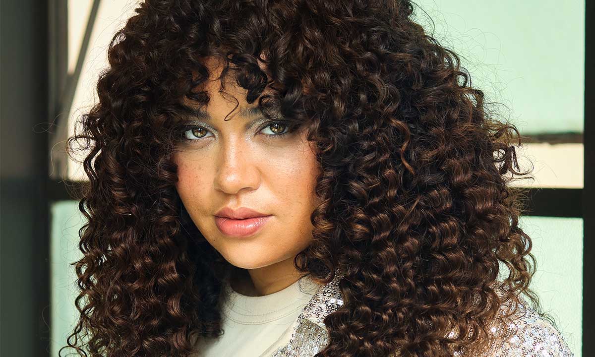 A person with long, dark curly hair styled in a festive holiday hairstyle and light eyes looks directly at the camera, standing indoors near a window.