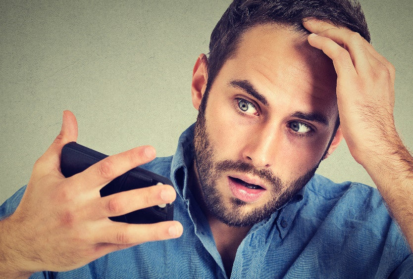 A man with a beard and blue shirt looks shocked while holding a phone in one hand and touching his hair with the other, perhaps searching for hair fibers or hair loss concealers as he stares at the phone screen, wide-eyed and mouth slightly open.