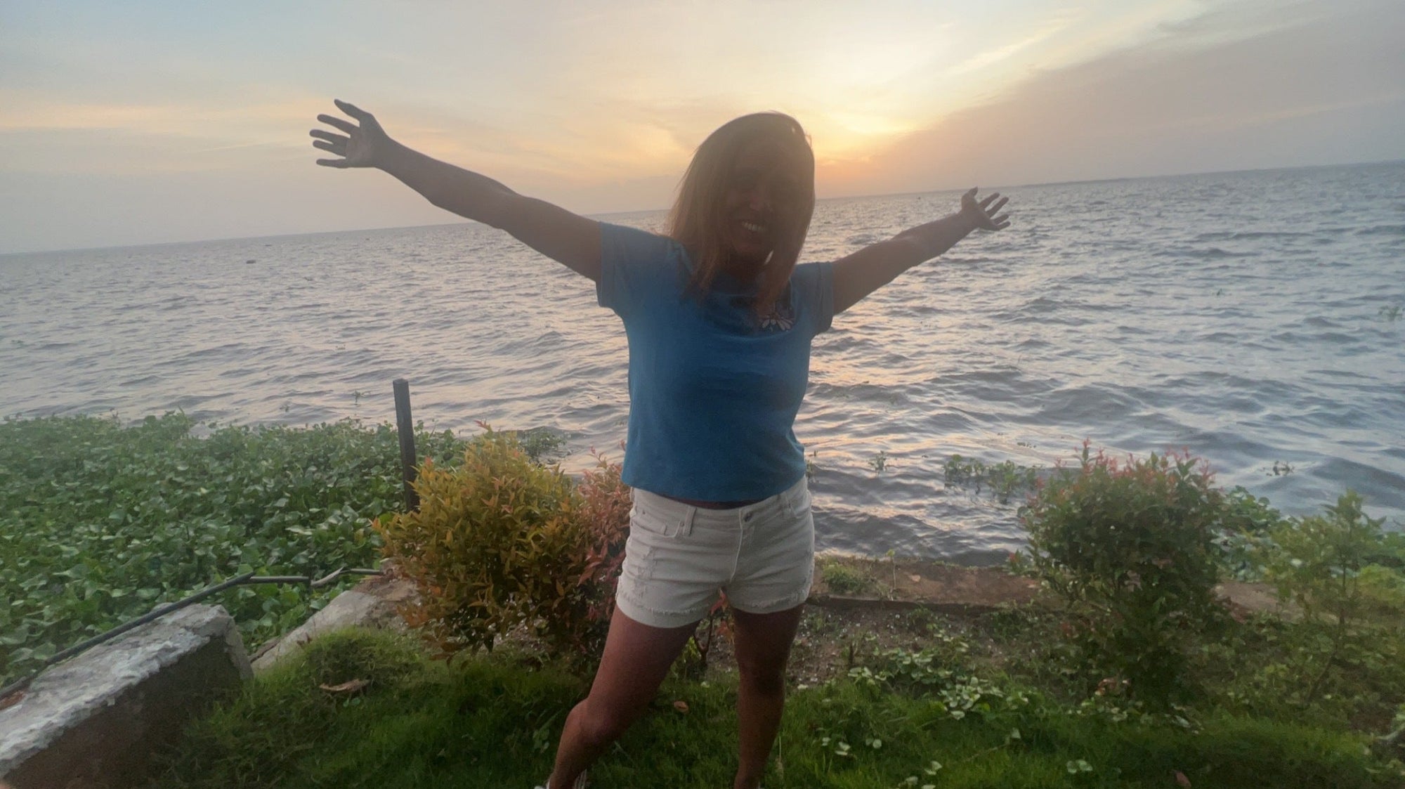 A person stands on grass by a lakeshore at sunset, arms outstretched and smiling, reflecting on their hair journey.