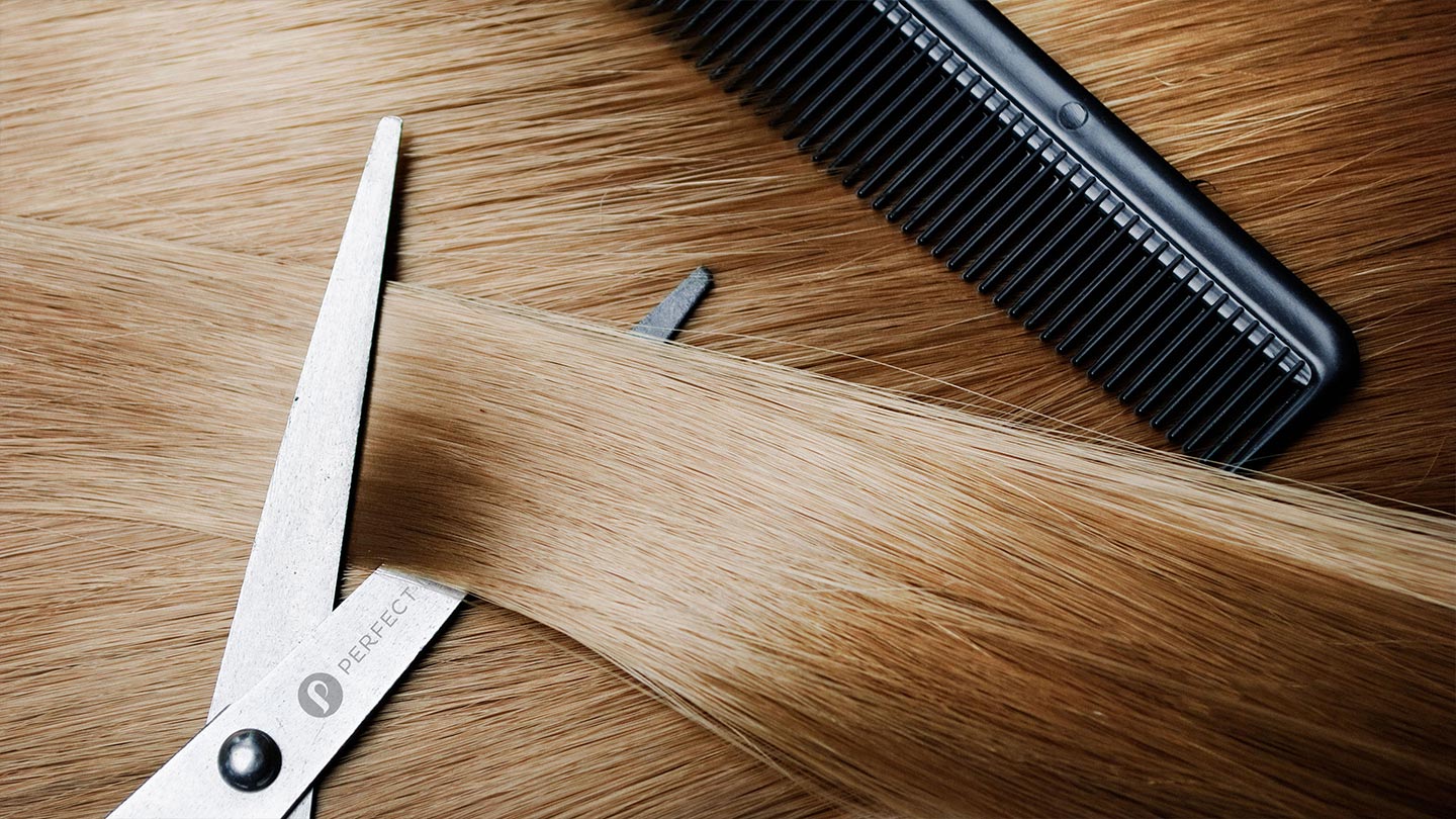 A pair of silver scissors cutting strands of light brown hair, with a black comb placed alongside the hair.