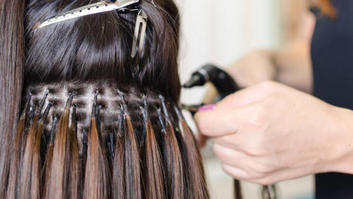 Close-up of a Salon Stylist attaching hair extensions to a person's natural hair with a small black tool; the hair is sectioned and clipped for easier Hair Extension application.