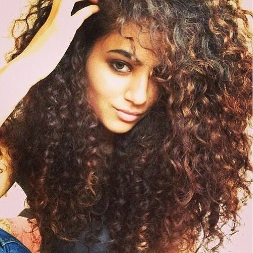 A person with voluminous, dark brown, curly hair looks at the camera, partially smiling, with one hand running through their hair.