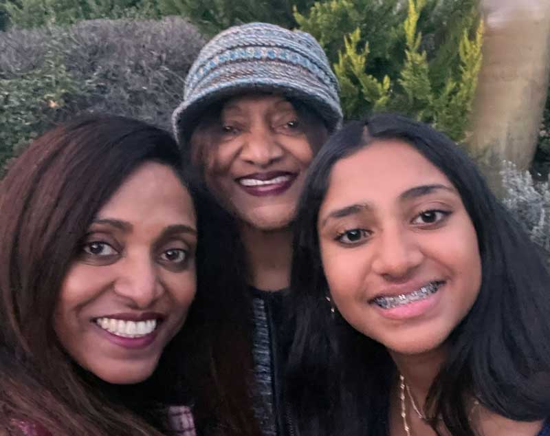 Three women smiling outdoors, including a girl with braces, a woman in a knit hat, and another woman, capture a joyful Mother’s Day memory.