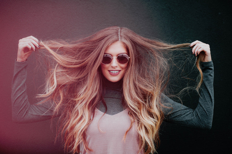 A woman with long, wavy brown hair, sunglasses, a dark long-sleeve top, and a light vest stands against a dark background, smiling and holding her hair out to the sides to show how tape-in extensions blend seamlessly.