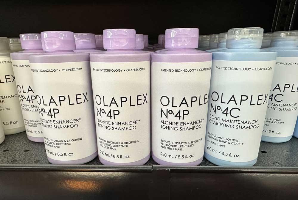 Several bottles of Olaplex shampoo are displayed on a shelf, including No. 4P Blonde Enhancer Toning Shampoo in lavender packaging and No. 4C Blonde Maintenance Clarifying Shampoo in light blue.