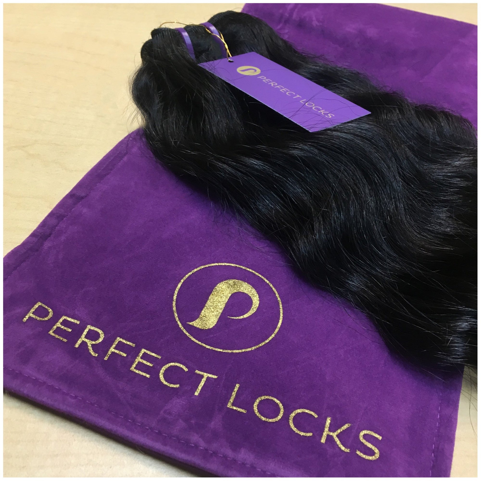 A bundle of wavy black hair extensions with a purple tag rests on a purple velvet bag labeled “Perfect Locks” in gold lettering, an elegant hair extension storage solution on a light wood surface.