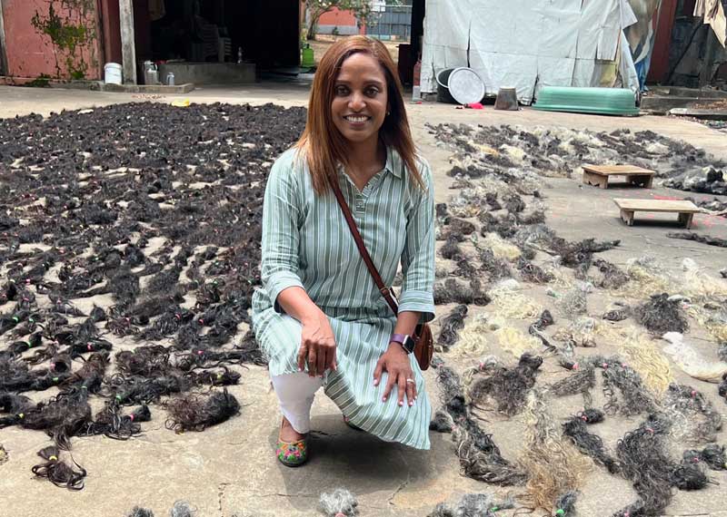 A woman in a striped dress kneels outdoors on a concrete surface covered with piles of cut hair, highlighting the hair journey, with sheds and equipment visible in the background.