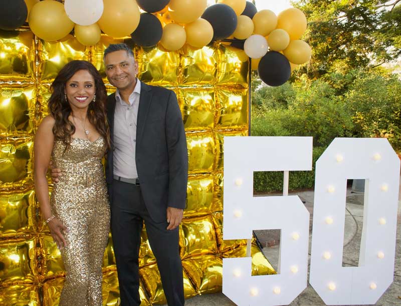 A smiling couple stands in front of a gold balloon backdrop and large illuminated 50 sign, celebrating milestones at an outdoor event.