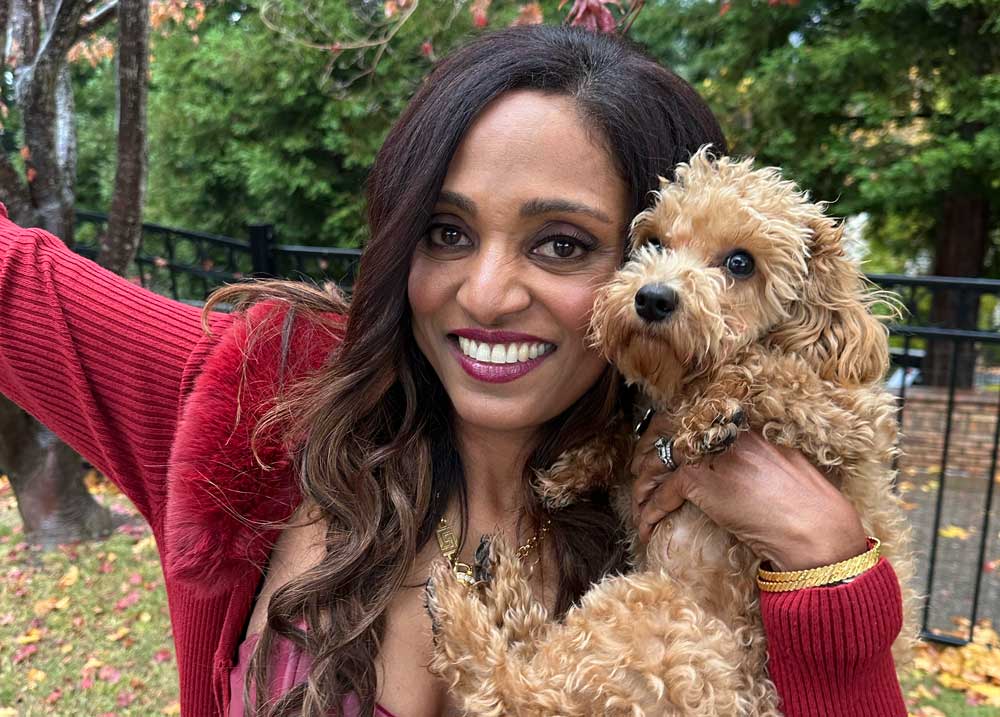 A smiling woman with long dark hair holds a small, curly-haired dog in her arms outdoors, with trees and a black fence in the background.