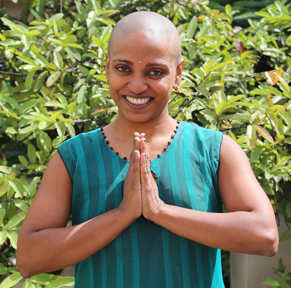 A smiling person with a shaved head stands outdoors in front of green foliage, wearing a sleeveless teal top and holding their hands together in a namaste gesture, perhaps reflecting on their hair restoration journey at a local hair clinic.