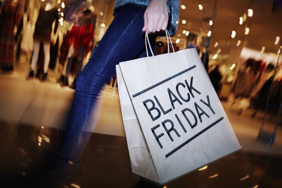 A person holding a shopping bag with BLACK FRIDAY printed on it stands in a brightly lit store, surrounded by blurred shoppers and displays, perfectly capturing the excitement of Black Friday shopping, just like Priyanka’s thoughts on great deals.