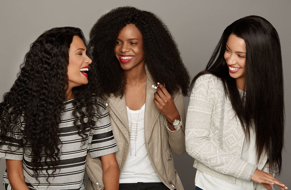 Three women with different hairstyles, curly, voluminous natural, and straight, smile and laugh together against a plain background, showing a vibrant hair comparison while wearing casual, stylish clothing in black, white, and beige tones.