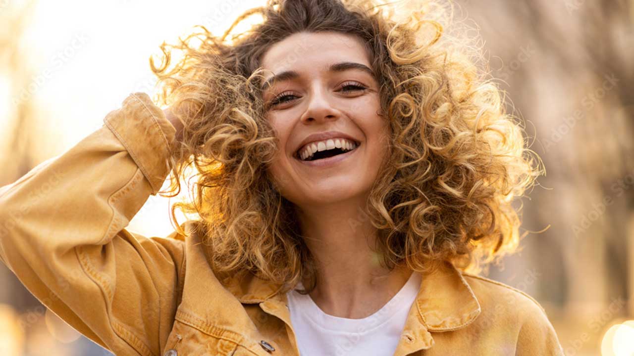 A young woman with curly, natural hair smiles brightly, wearing a yellow jacket and white shirt, standing outdoors in warm sunlight and embracing her unique hair texture.