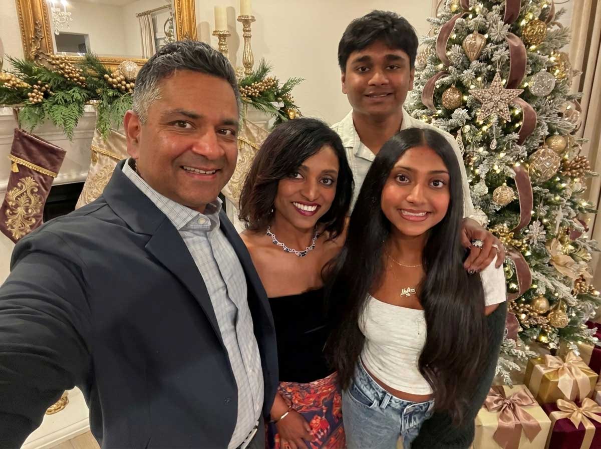 A family of four poses and smiles in front of a decorated Christmas tree with gold ornaments and wrapped presents, sharing gratitude and holiday wishes in their festive, warmly lit living room.